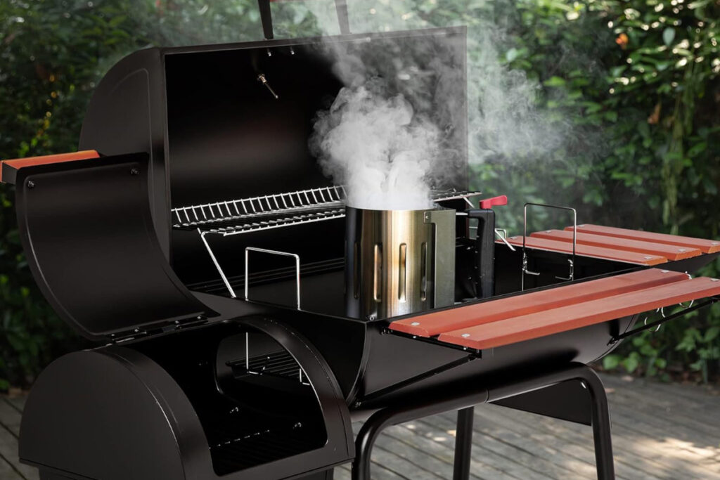 "Royal Gourmet CC1830W Charcoal Grill with smoke rising from a chimney starter, featuring a sleek black design and wooden side shelves, set in a lush outdoor environment."